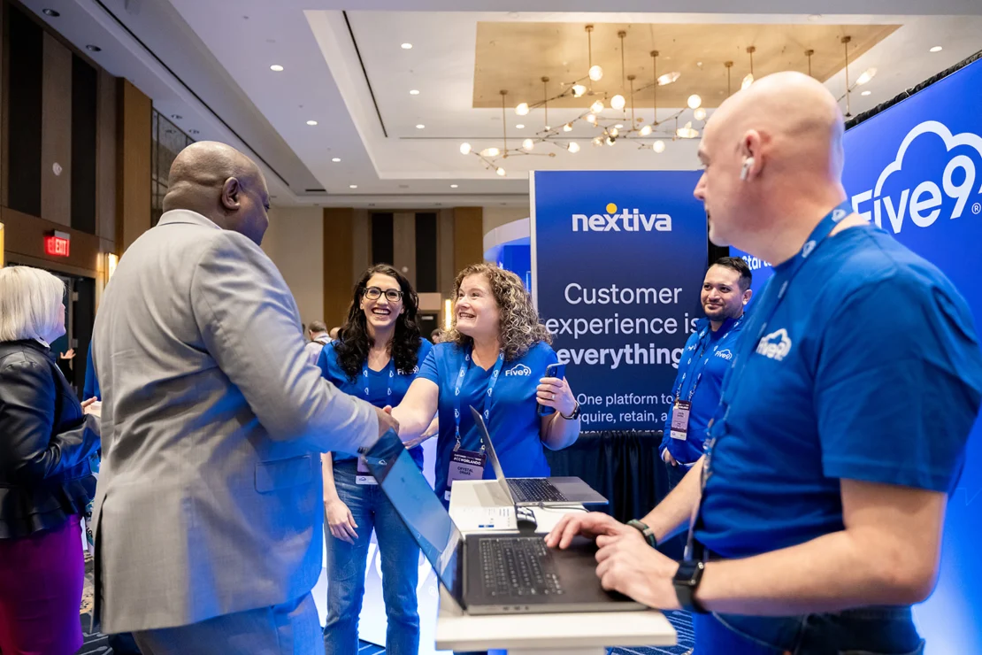 A group of people at a trade show booth with "Five9" and "Nextiva" branding, engaging in conversation and smiling; one person is using a laptop.
