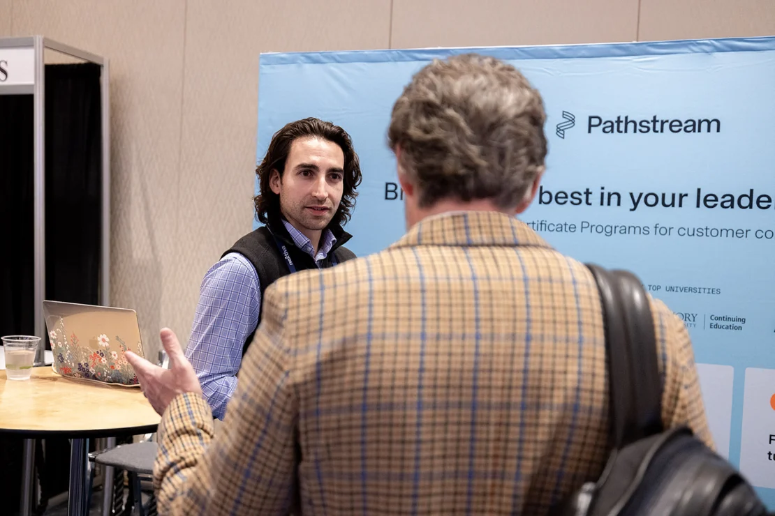 Two men talk at a Pathstream booth; one faces the camera, the other has his back to it. A laptop and cup are on a nearby table.