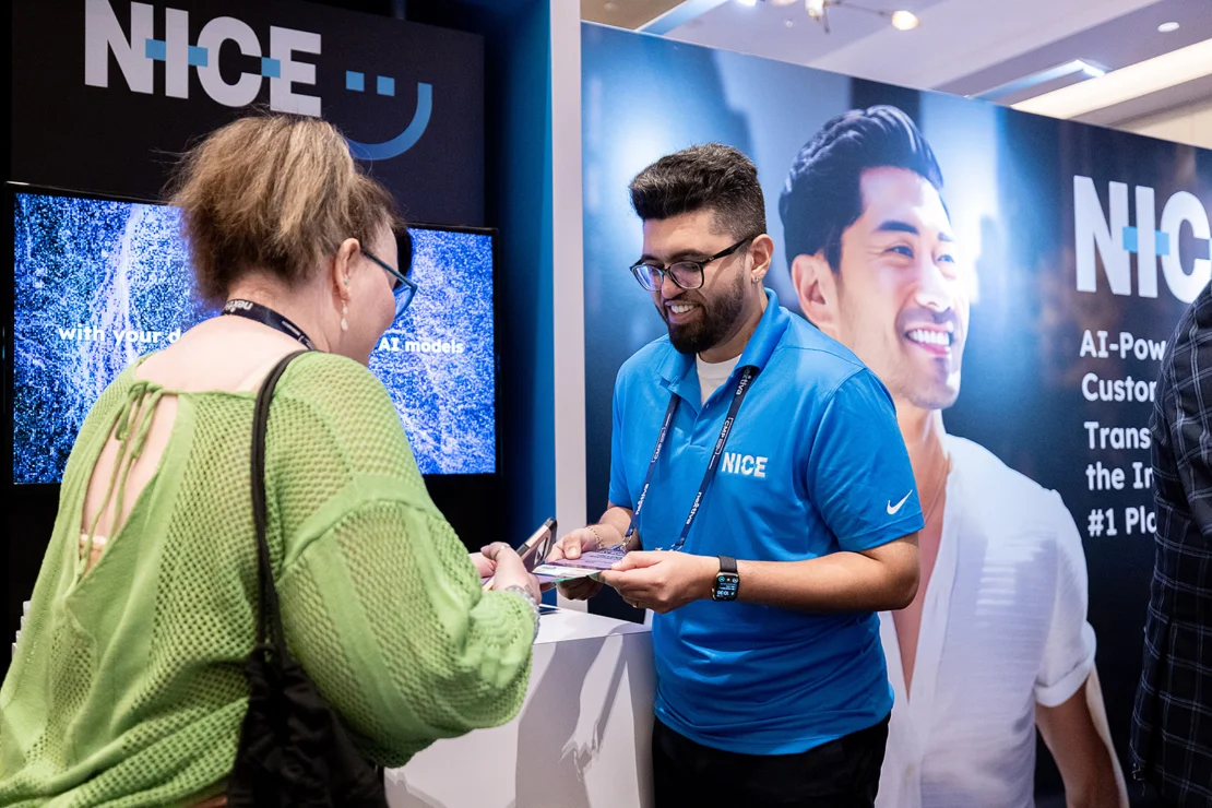 A man in a blue NICE shirt assists a woman at an exhibition booth with NICE branding and promotional displays in the background.