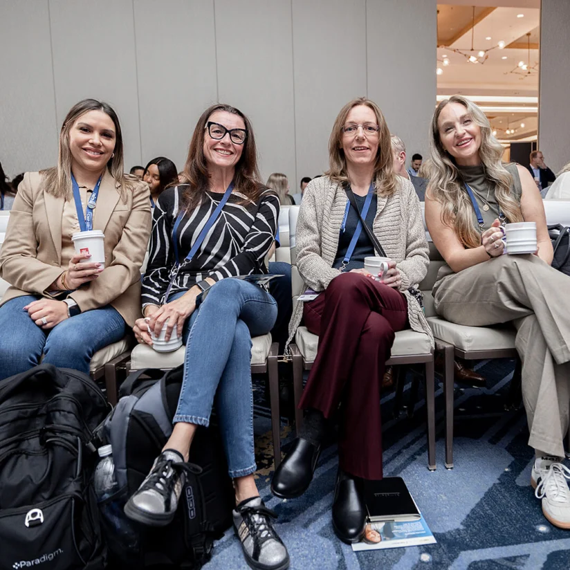 Four women sit in a row of white chairs at an indoor event, holding drinks and smiling at the camera, with other attendees visible in the background.