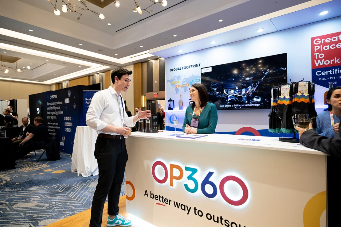 Two people talk at an OP360 booth in a conference hall, with informational displays and promotional materials visible on the counter and walls.