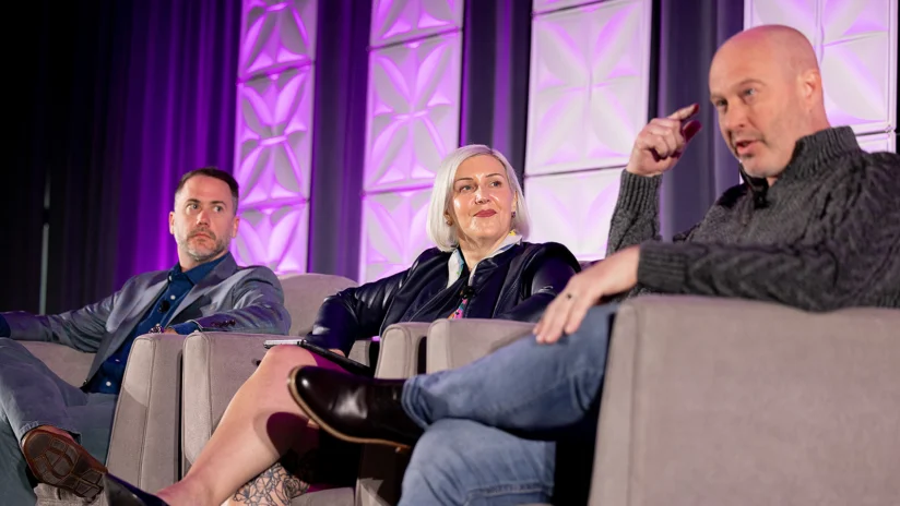 Three people sit on stage in armchairs, engaged in a panel discussion with purple geometric lighting in the background.