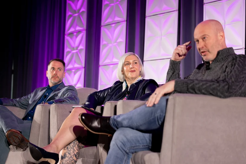 Three people sit on stage in armchairs, engaged in a panel discussion with purple geometric lighting in the background.
