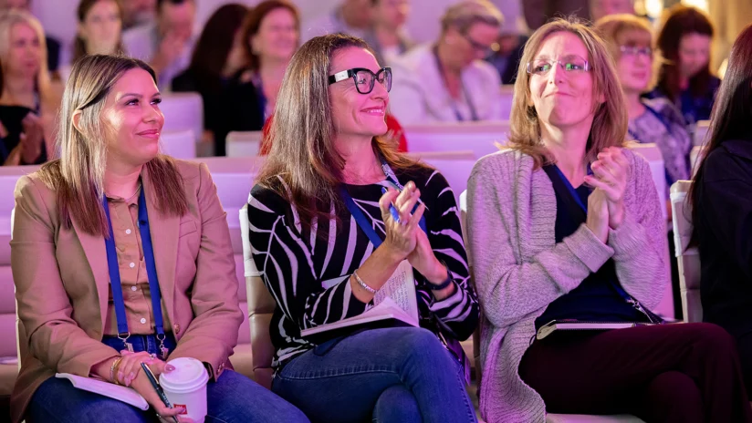 Three women sitting in a crowd, smiling and clapping, with notebooks and a coffee cup, at what appears to be a conference or seminar.