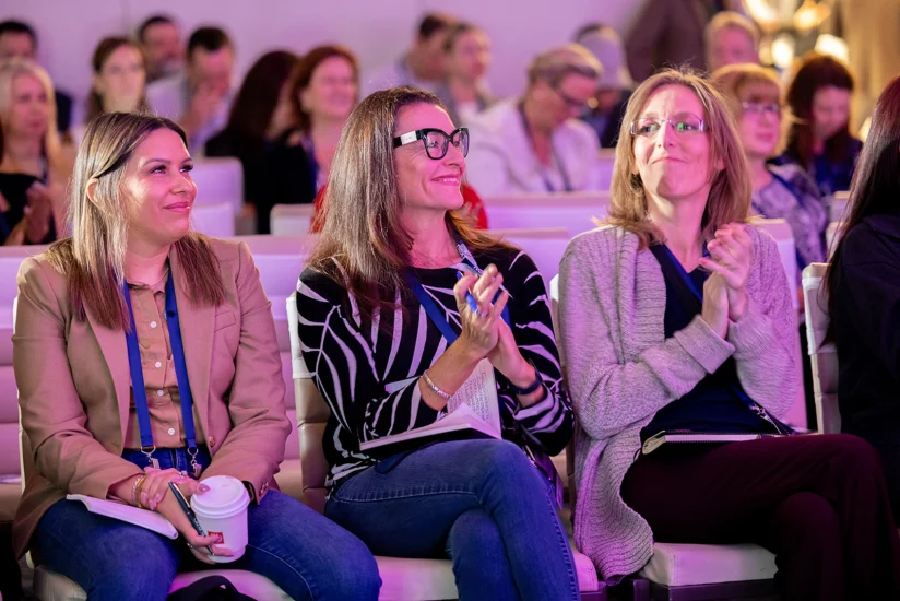 Three women sitting in a crowd, smiling and clapping, with notebooks and a coffee cup, at what appears to be a conference or seminar.