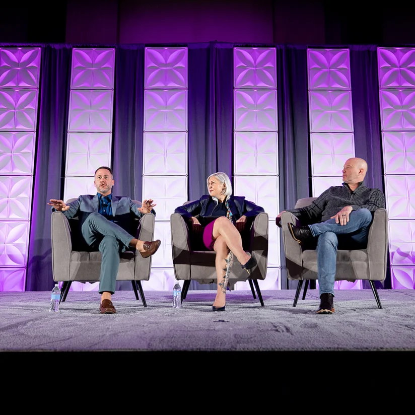 Three people sit and speak on a stage with geometric purple-lit panels behind them, participating in a panel discussion.