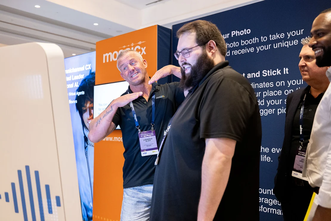Four men stand at a photo booth during a conference; one man poses playfully while the others smile and look at the screen.