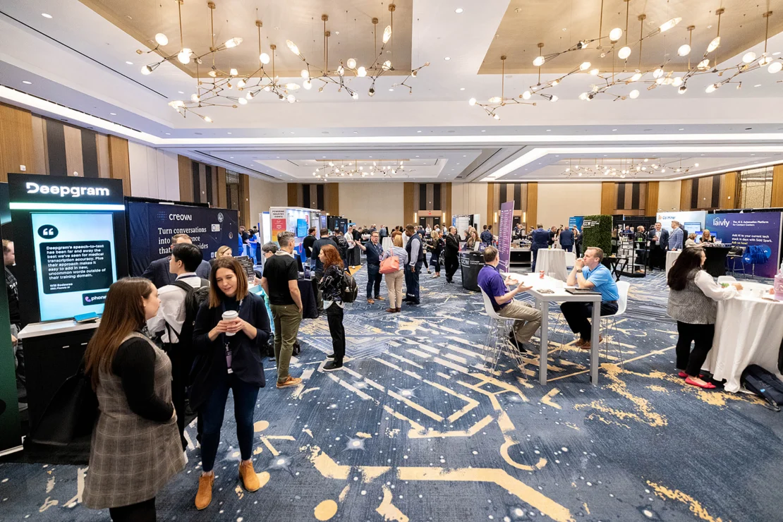 People attending a tech conference mingle and visit various company booths in a large, modern convention hall with blue carpeting and bright lighting.