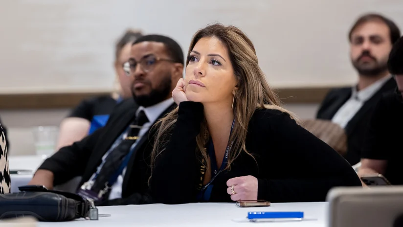 A woman sits at a table with others, resting her chin on her hand and looking forward attentively during a meeting or conference.