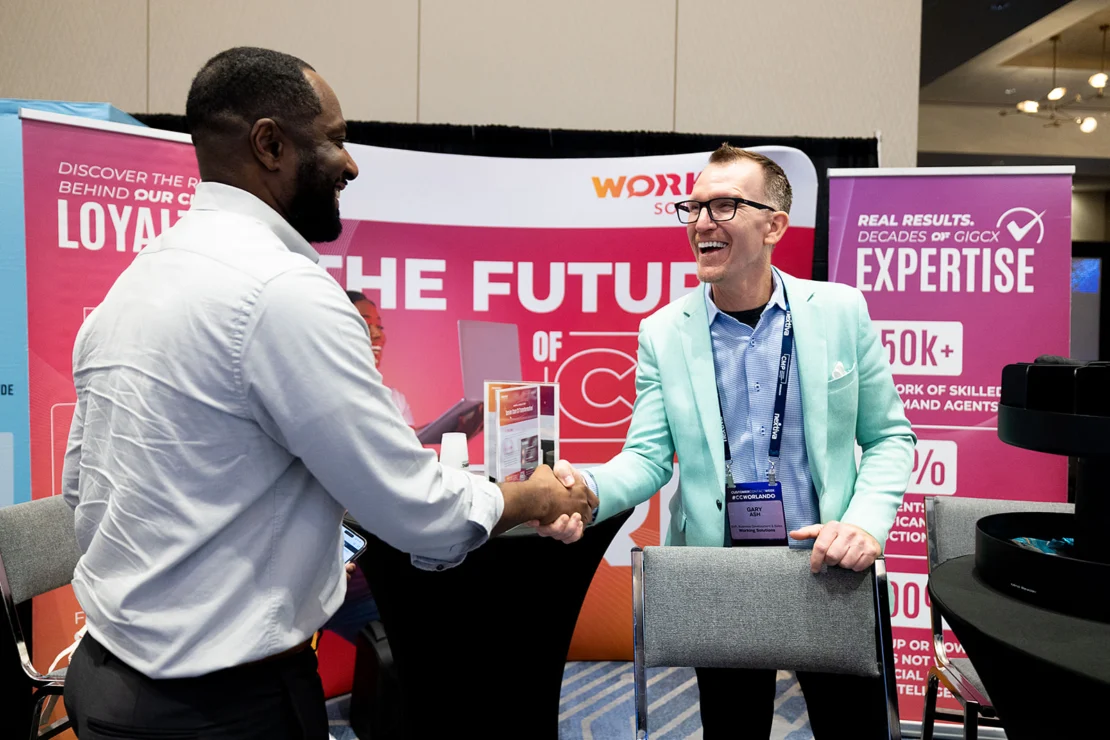Two men in business attire smile and shake hands at a conference booth with promotional banners in the background.