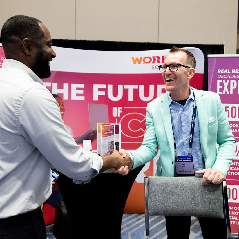 Two men in business attire smile and shake hands at a conference booth with promotional banners in the background.
