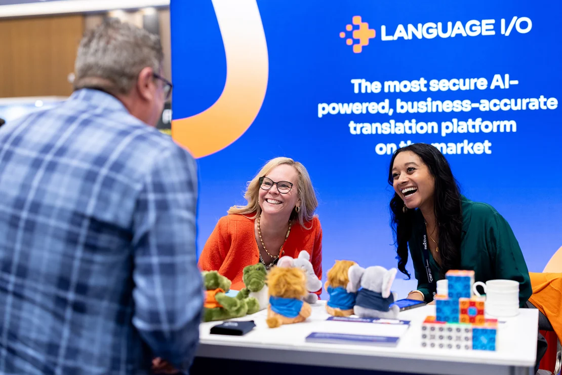 Two people sit and smile at a trade show booth for Language I/O, featuring stuffed animals and cube puzzles on the table. A man stands across from them, engaged in conversation.