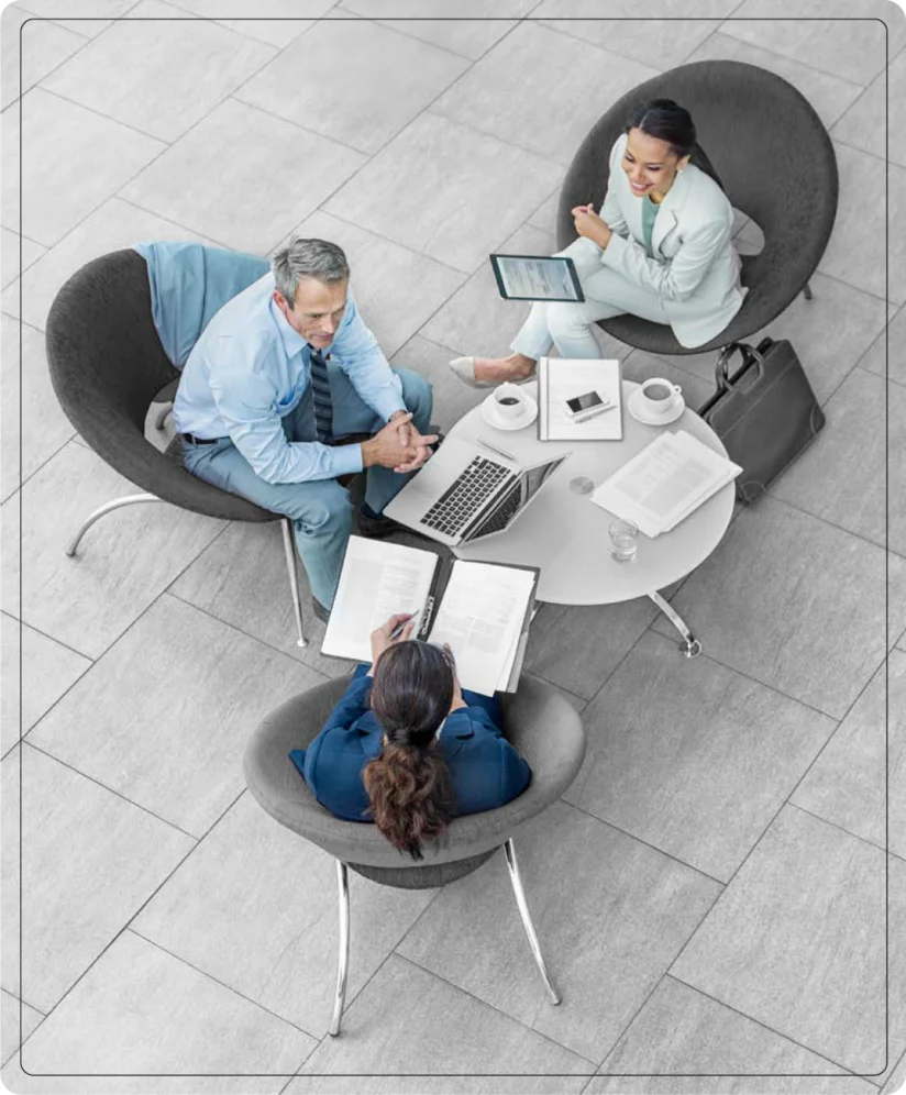 Three people in business attire sit around a small table with laptops, documents, and coffee, engaged in a meeting on a tiled floor.