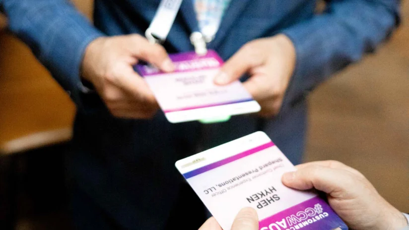 Two people in business attire hold and look at conference name badges while standing indoors.