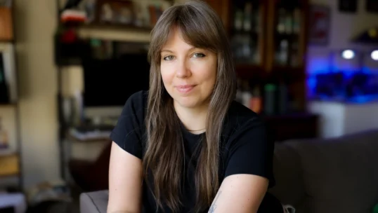 A woman with straight brown hair and bangs sits on a couch in a room with shelves and cabinets in the background, looking at the camera with a neutral expression.