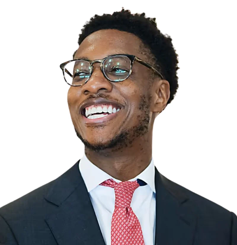 A man in a dark suit, white shirt, and red patterned tie is smiling and looking to the side. He is wearing glasses and has short curly hair. The background is white.