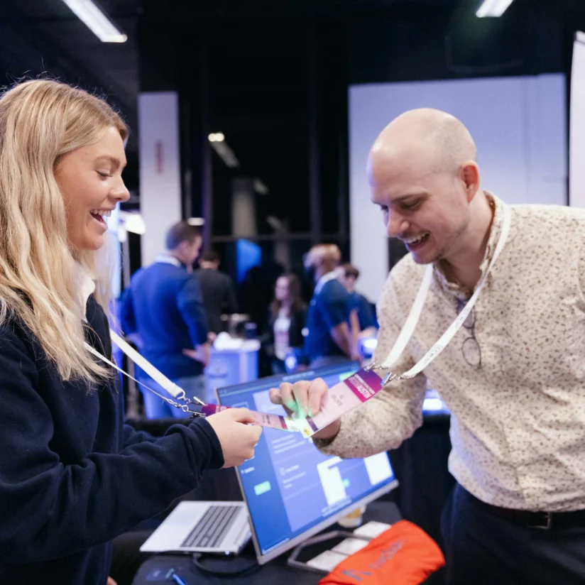 A woman hands a badge to a smiling man at a booth during a business event or conference, with banners and computers in the background.