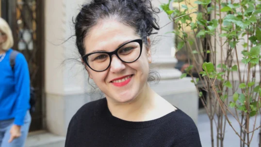 A woman with curly dark hair, black glasses, and a black top stands smiling outdoors near a plant, with city buildings in the background.