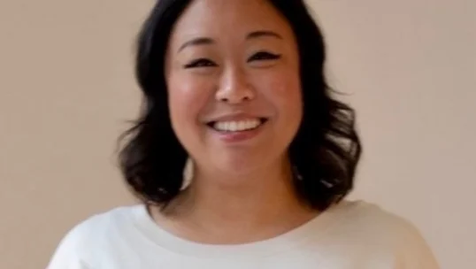 A woman with medium-length dark hair, wearing a white top, smiles while standing in front of a plain light-colored background.