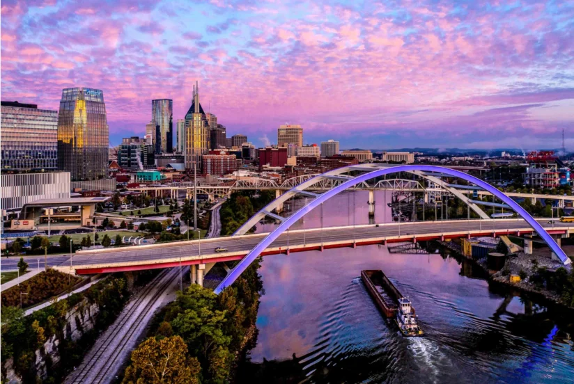 A barge travels under an arched bridge on a river, with a modern city skyline and a colorful sunset sky in the background.