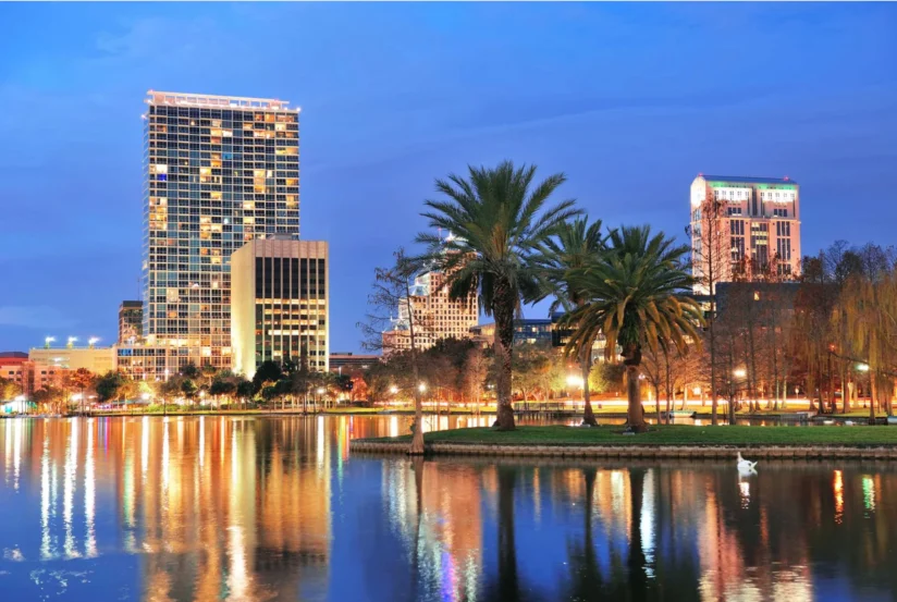 City skyline with modern high-rise buildings, palm trees, and a lake reflecting the illuminated cityscape at dusk.