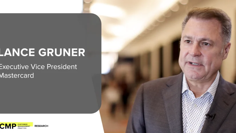 Lance Gruner, Executive Vice President at Mastercard, stands in a hallway and speaks during a professional event. A name and title banner appears on the left.