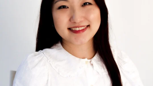 A woman with long dark hair wearing a white blouse sits in front of a plain white background, smiling at the camera.