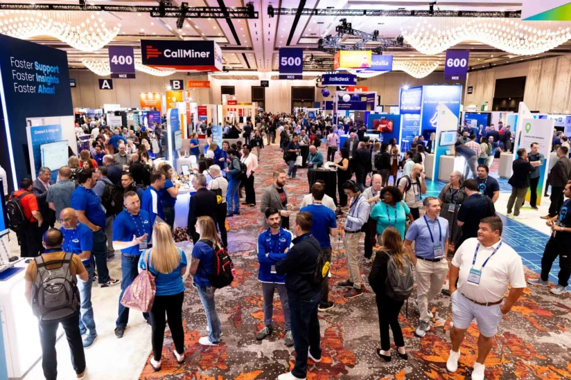 Large crowd of people attending a technology conference, walking and talking among various exhibition booths and displays in a spacious, well-lit convention hall.