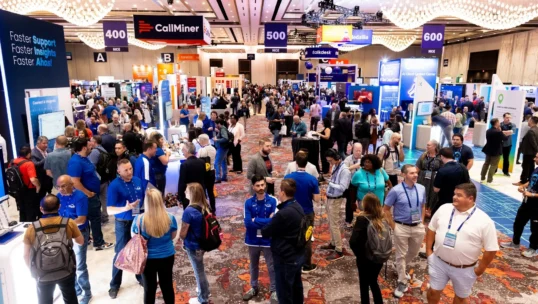 Crowds of attendees walk and interact at a busy technology conference expo, surrounded by booths and banners from various companies in a large, well-lit convention hall.
