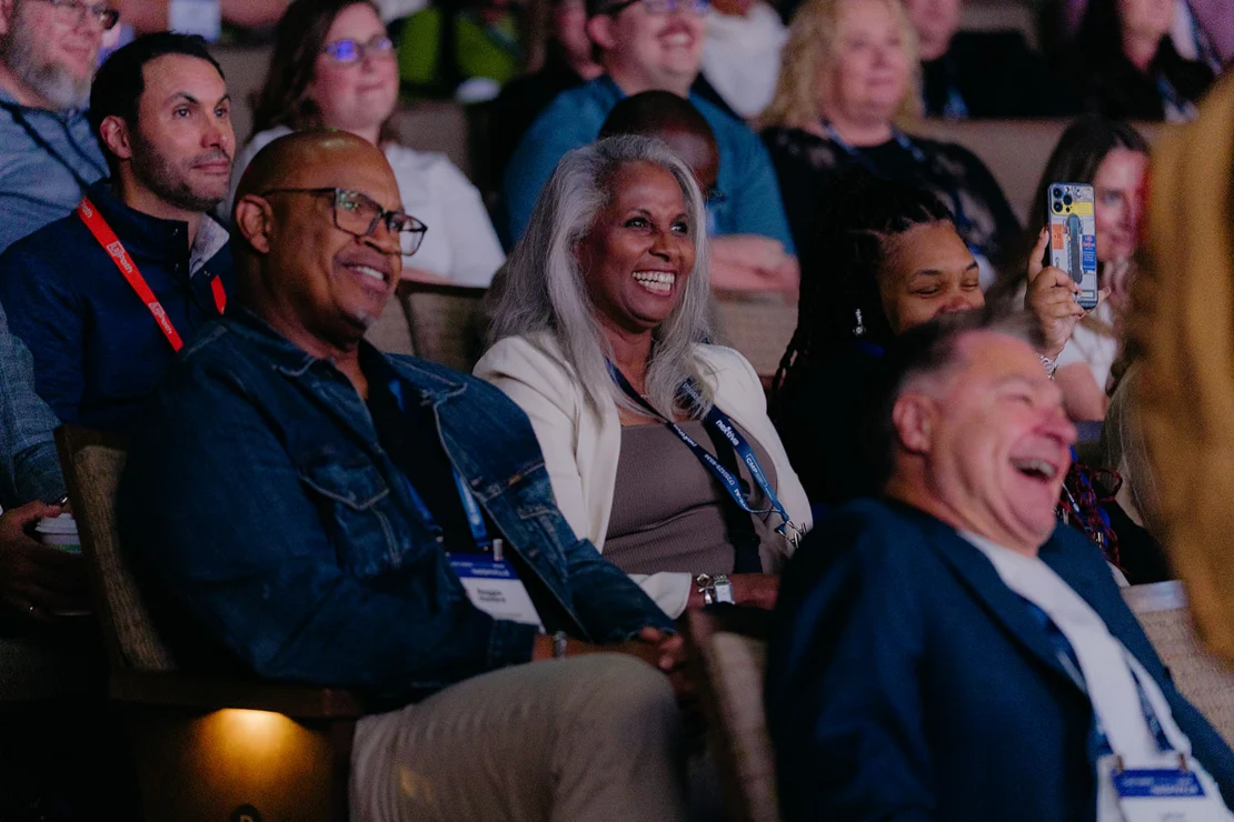 A group of people sit in an audience, smiling and laughing while watching an event or presentation.