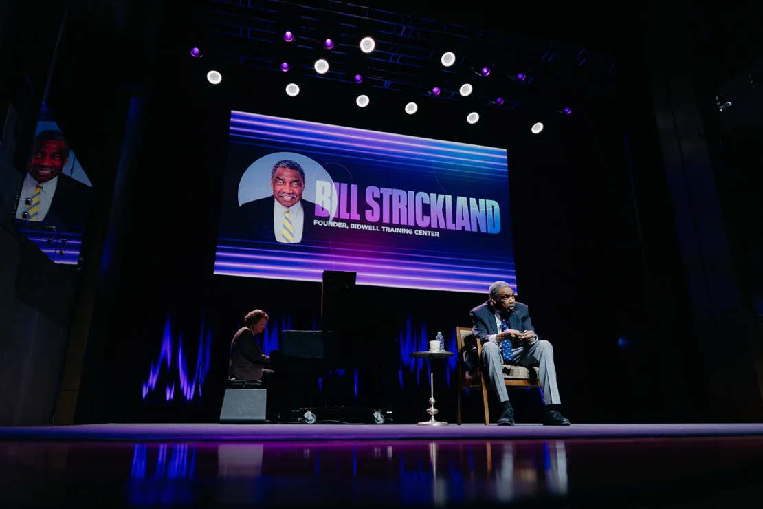 A man sits on stage near a pianist, with a large screen behind displaying “Bill Strickland, Founder, Bidwell Training Center” and his photo.