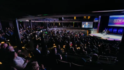 A large audience sits in a darkened auditorium watching a speaker on stage at a conference, with the event being displayed on a screen above.