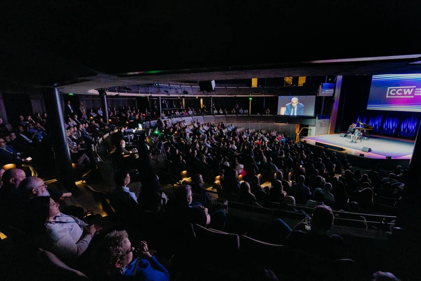 A large audience sits in a darkened auditorium watching a speaker on stage at a conference, with the event being displayed on a screen above.