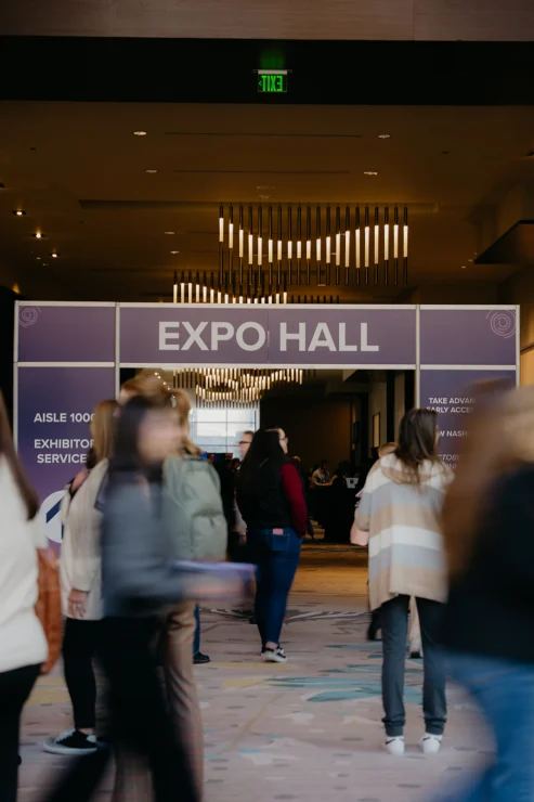 People walk toward and enter an expo hall through large doors under a sign reading "Expo Hall" in a convention center setting.