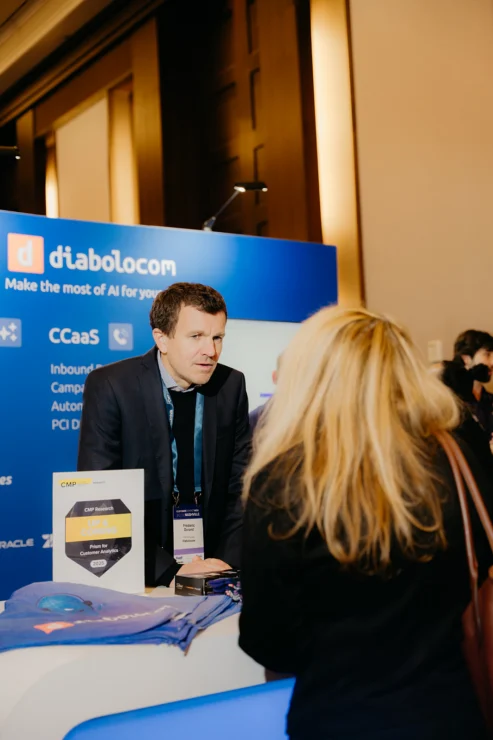 A man stands behind a Diabolocom booth at a conference, speaking to an attendee. Booth displays brochures, branded items, and a sign about an award.