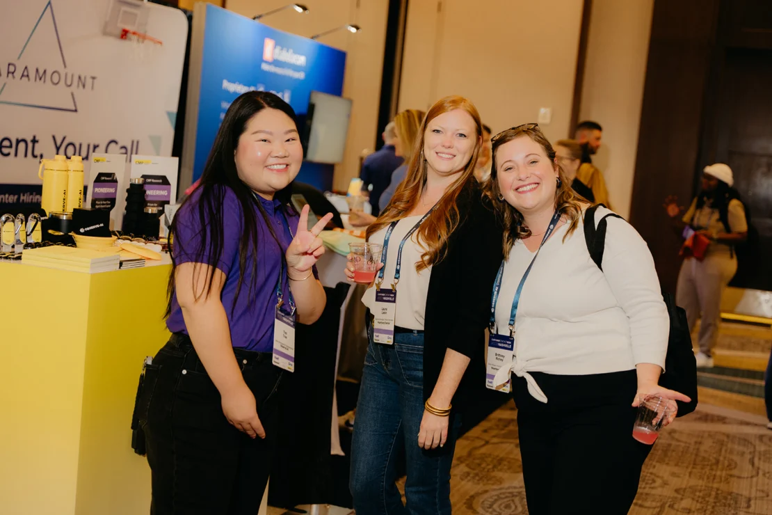 Three women with conference badges smile for a photo at an indoor event, standing near a yellow table with promotional items and displays in the background.