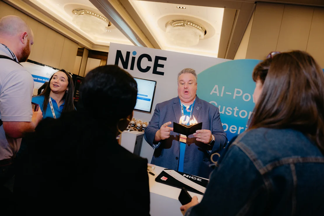 A man stands at a NiCE booth at an event, holding a booklet and speaking to a small group of people. AI-powered customer experience signage is visible behind him.