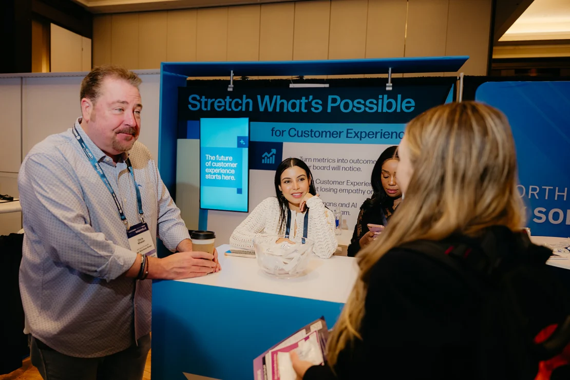A man talks to three women at a booth with a sign reading “Stretch What’s Possible for Customer Experience” at a convention or expo.