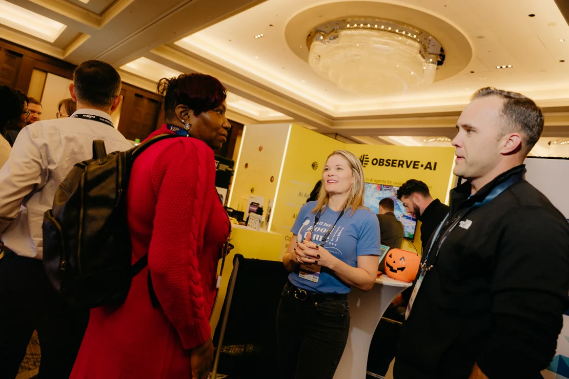People converse at a conference booth with "Observe.AI" branding; one person holds a pumpkin bucket, and a chandelier hangs from the ceiling above.