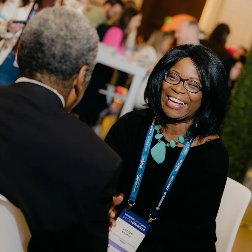 A woman wearing glasses and a name badge smiles while talking to a man at a busy indoor event.