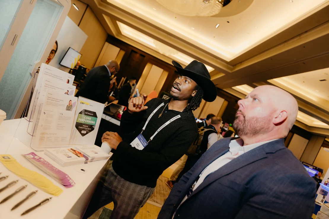Two men stand at a conference booth, one wearing a black hat and gesturing while the other in a suit listens attentively; brochures and display materials are visible on the table.