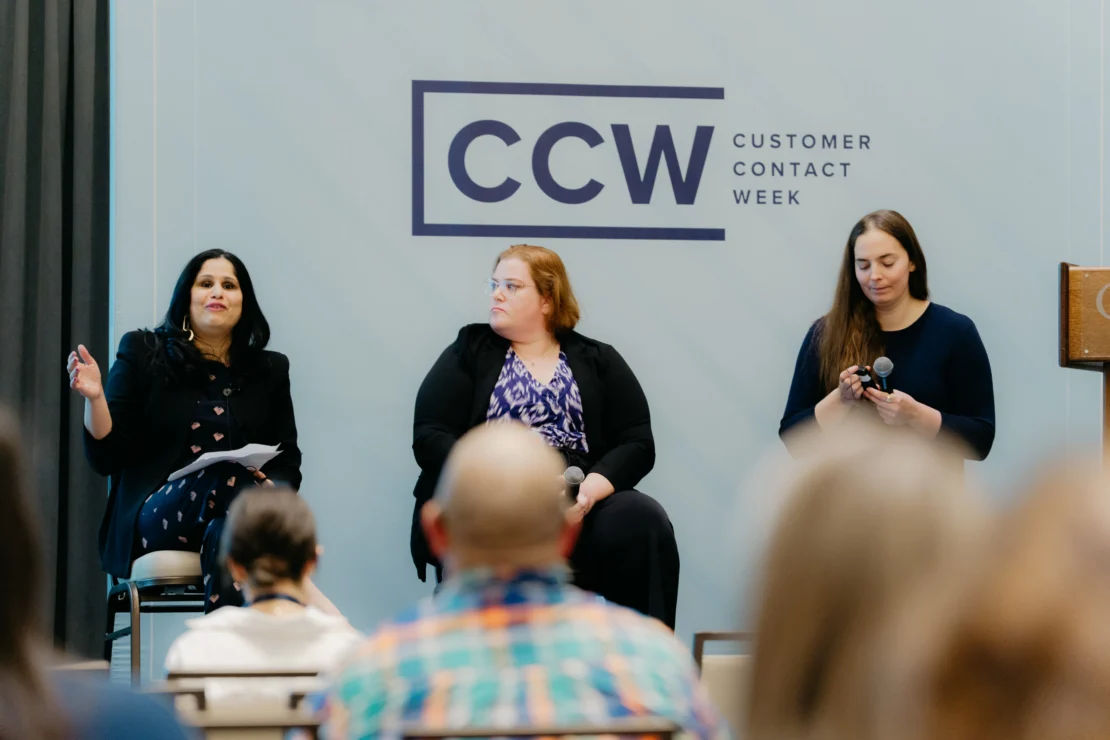 Three women sit on a panel discussion at Customer Contact Week, with one speaking, one listening, and one holding a microphone. Audience members are visible in the foreground.