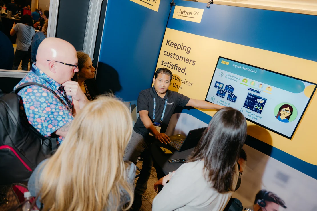 A presenter explains a customer service software display to a small group at a Jabra GN booth, with a screen showing an Agent Assistant interface.