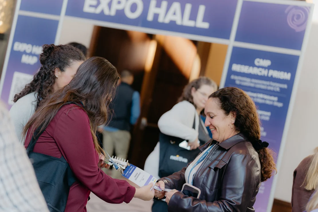 Several people interact and exchange materials near the entrance to an expo hall, with a large sign labeled "EXPO HALL" visible in the background.