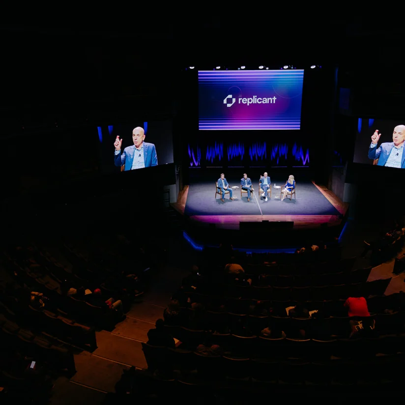 A panel of four people sits on stage in front of an audience, with "replicant" displayed on the screen behind them. The event is held in a dark theater.