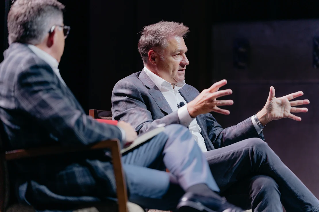 Two men in suits sit on stage having a discussion; one man gestures with his hands while speaking, and the other listens, holding a notebook.