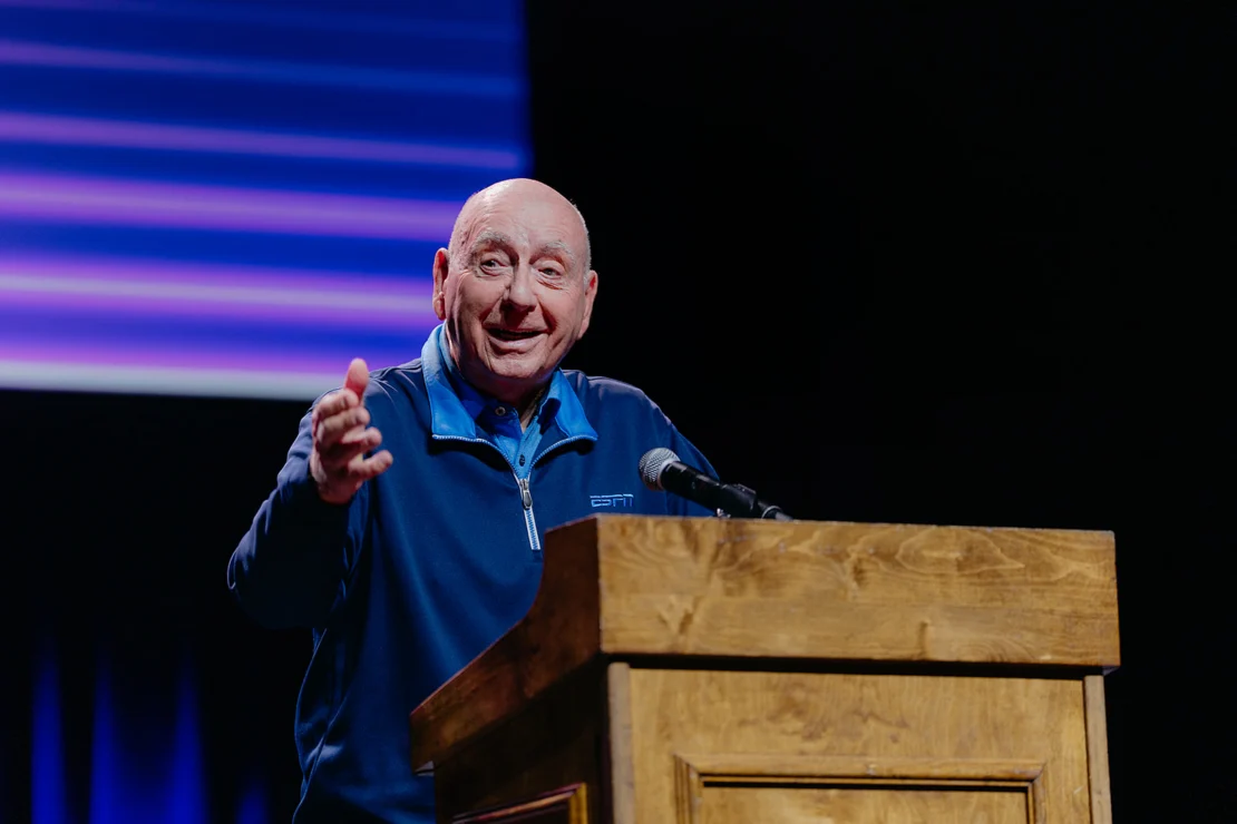 A man in a blue pullover speaks at a wooden podium on a stage with a dark background and blue-purple lighting.