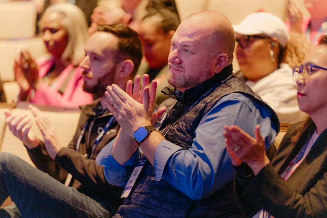 A group of people sit and clap during an indoor event or conference, focusing on a man in a blue shirt and vest in the foreground.