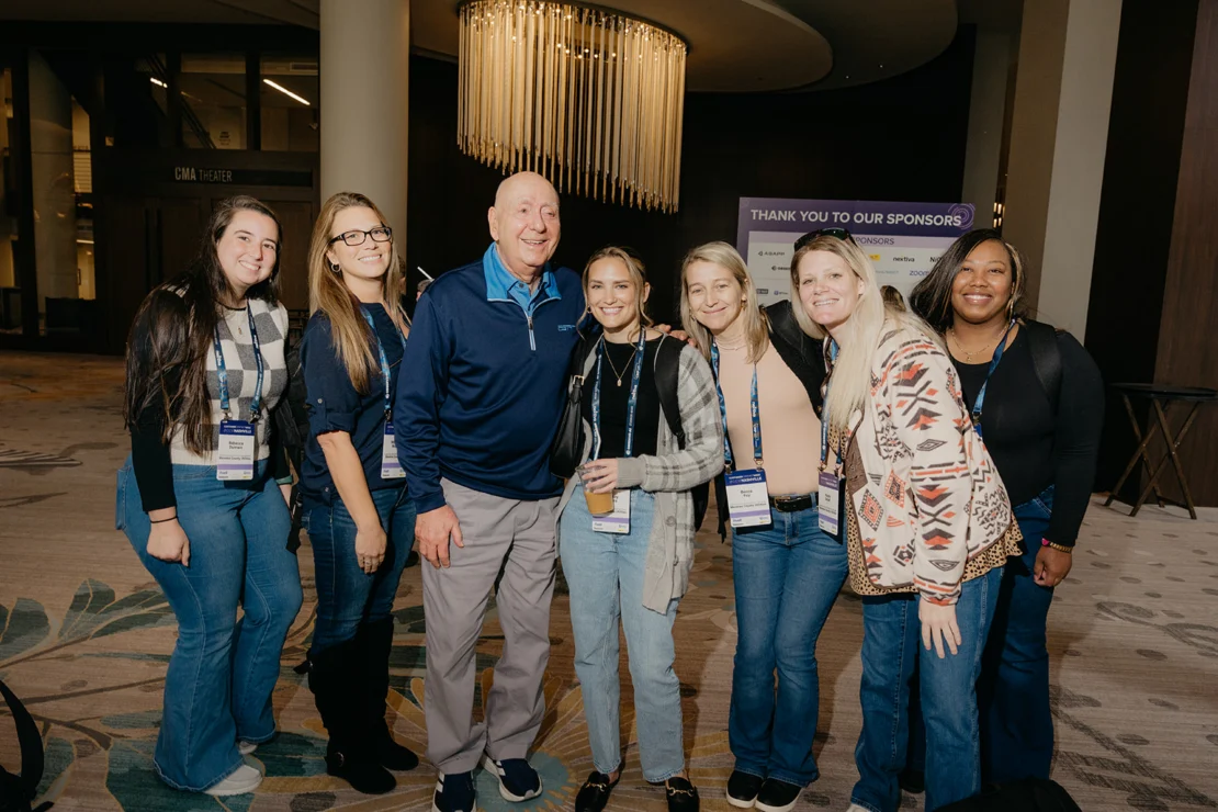 Seven people pose together indoors at an event, all wearing name badges. A “Thank You to Our Sponsors” sign is visible in the background.