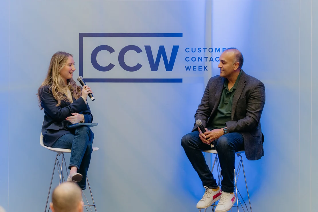 Two people sit on stools having a discussion with microphones in front of a CCW (Customer Contact Week) event backdrop.
